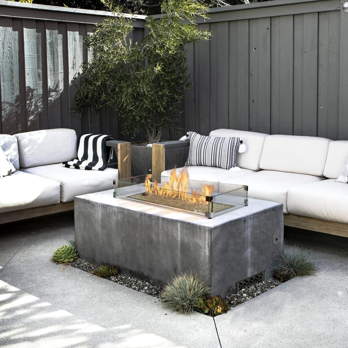 A concrete rectangular fire table with an active flame surrounded by a white outdoor sectional sofa on a modern backyard patio.