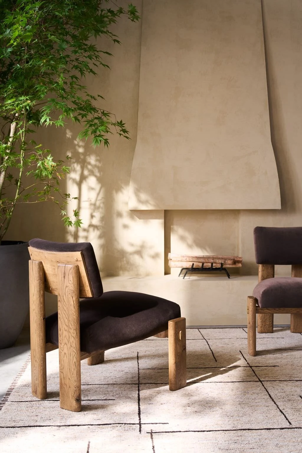Two natural oak frame accent chairs with dark charcoal upholstered seats arranged beside a large potted tree in front of a plaster fireplace surround with warm sunlight casting shadows.
