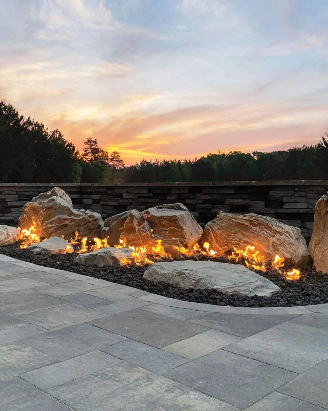 A custom linear fire pit with active flames on a waterfront deck at sunset, surrounded by outdoor seating.
