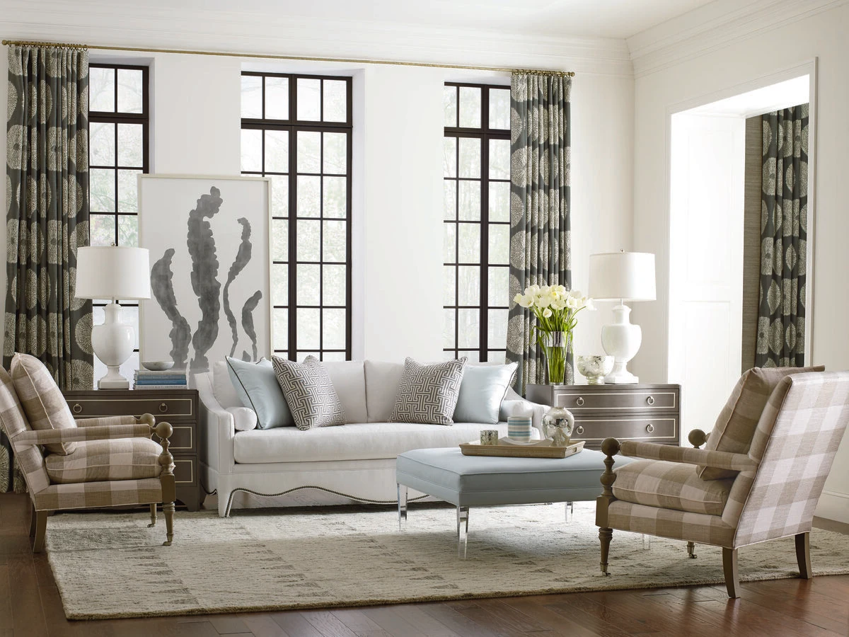 A bright transitional living room with a white upholstered sofa, neutral accent chairs, a glass coffee table, and floor-to-ceiling black steel windows with botanical print drapery.