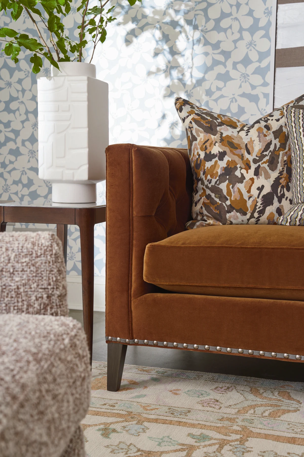 A close-up of a rust orange velvet accent chair with nailhead trim and a multicolored floral throw pillow, set against a blue botanical wallpaper backdrop.