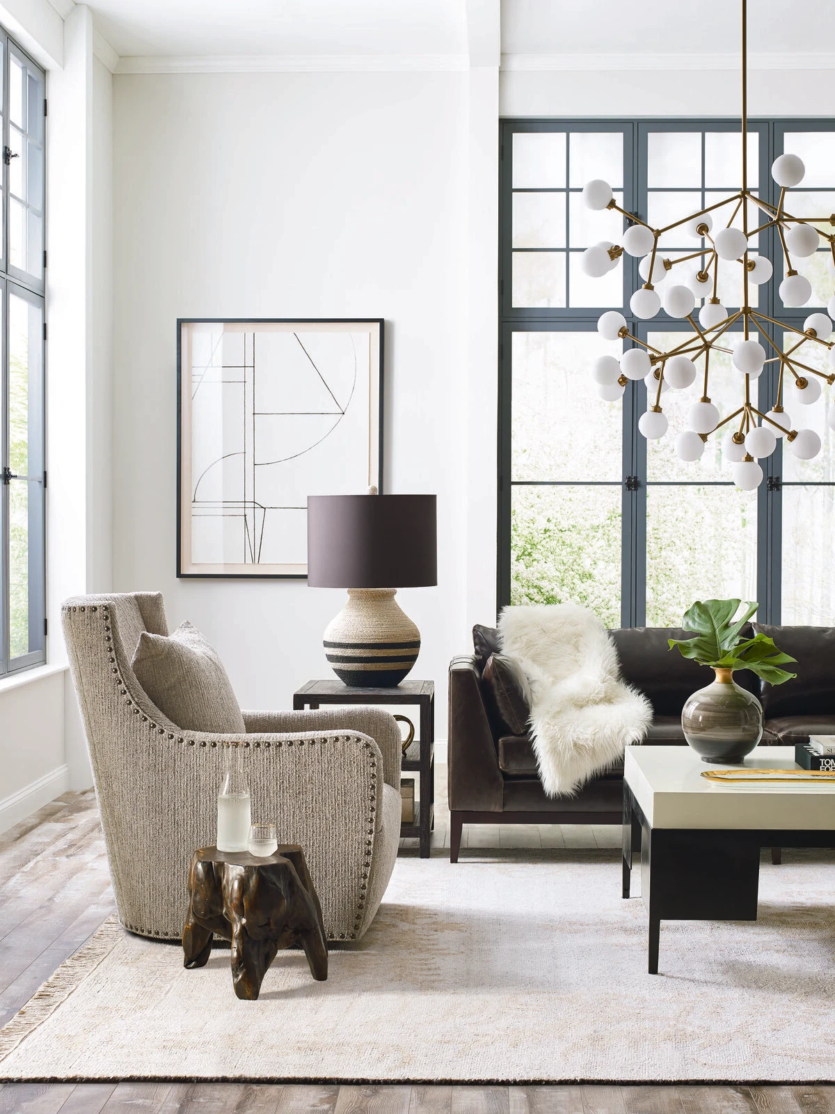 A contemporary living room with a gray nailhead accent chair, black leather sofa with a white fur throw, a modern brass globe chandelier, and black steel windows.