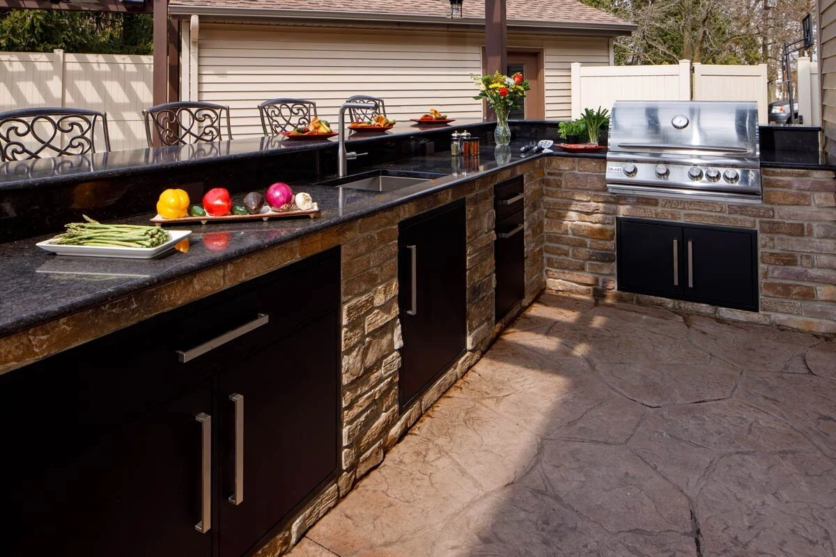 Custom outdoor kitchen with dark cabinetry, stainless steel grill, and brick accent wall. Shop outdoor kitchens near Spokane and North Idaho.