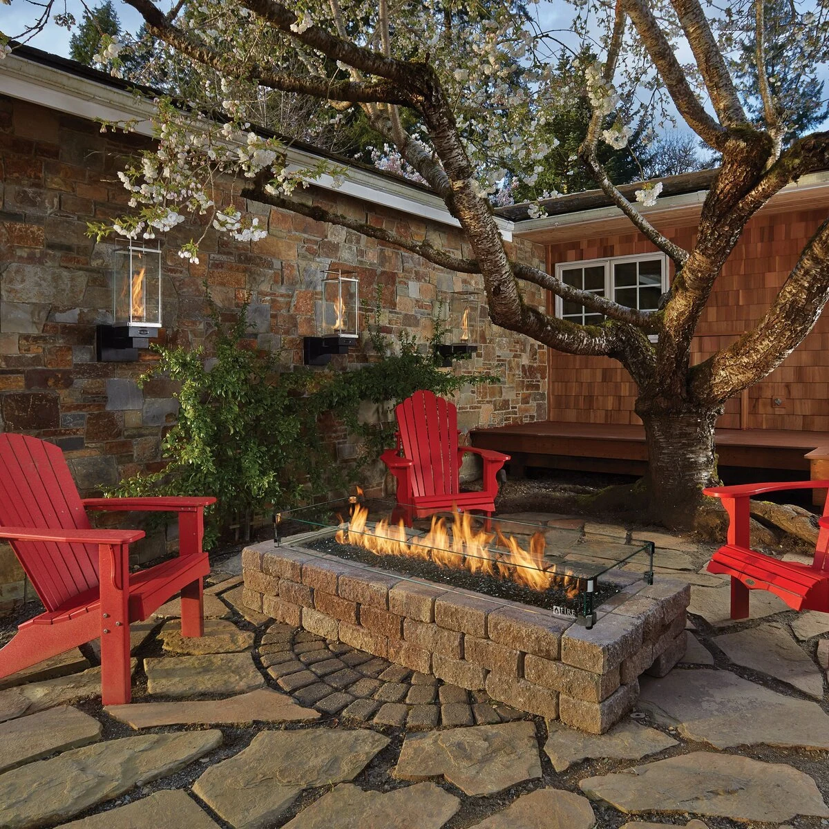 A stone fire pit with an active wood fire set into a rustic outdoor patio with red Adirondack chairs.
