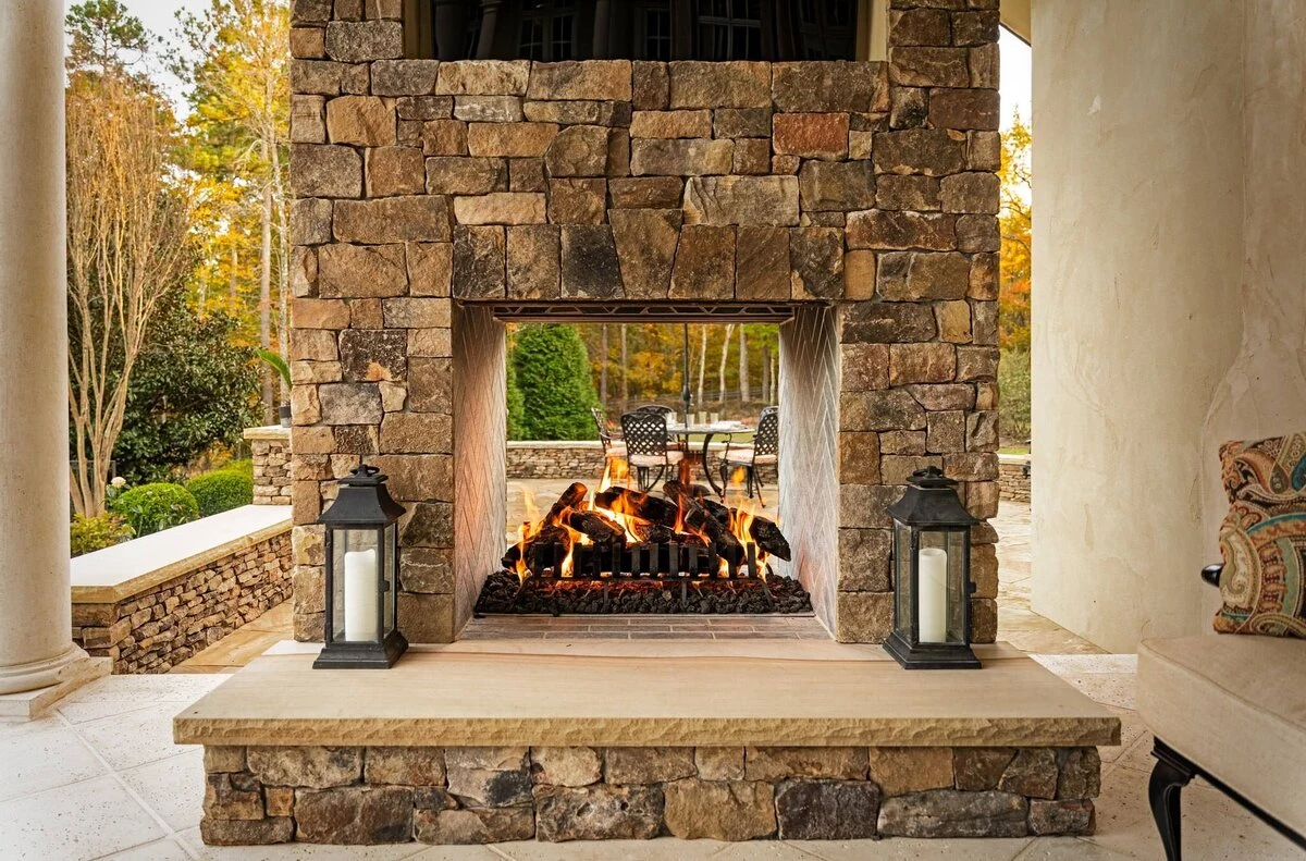 A custom stone outdoor fireplace with an active fire set into a floor-to-ceiling stone surround on a covered patio.