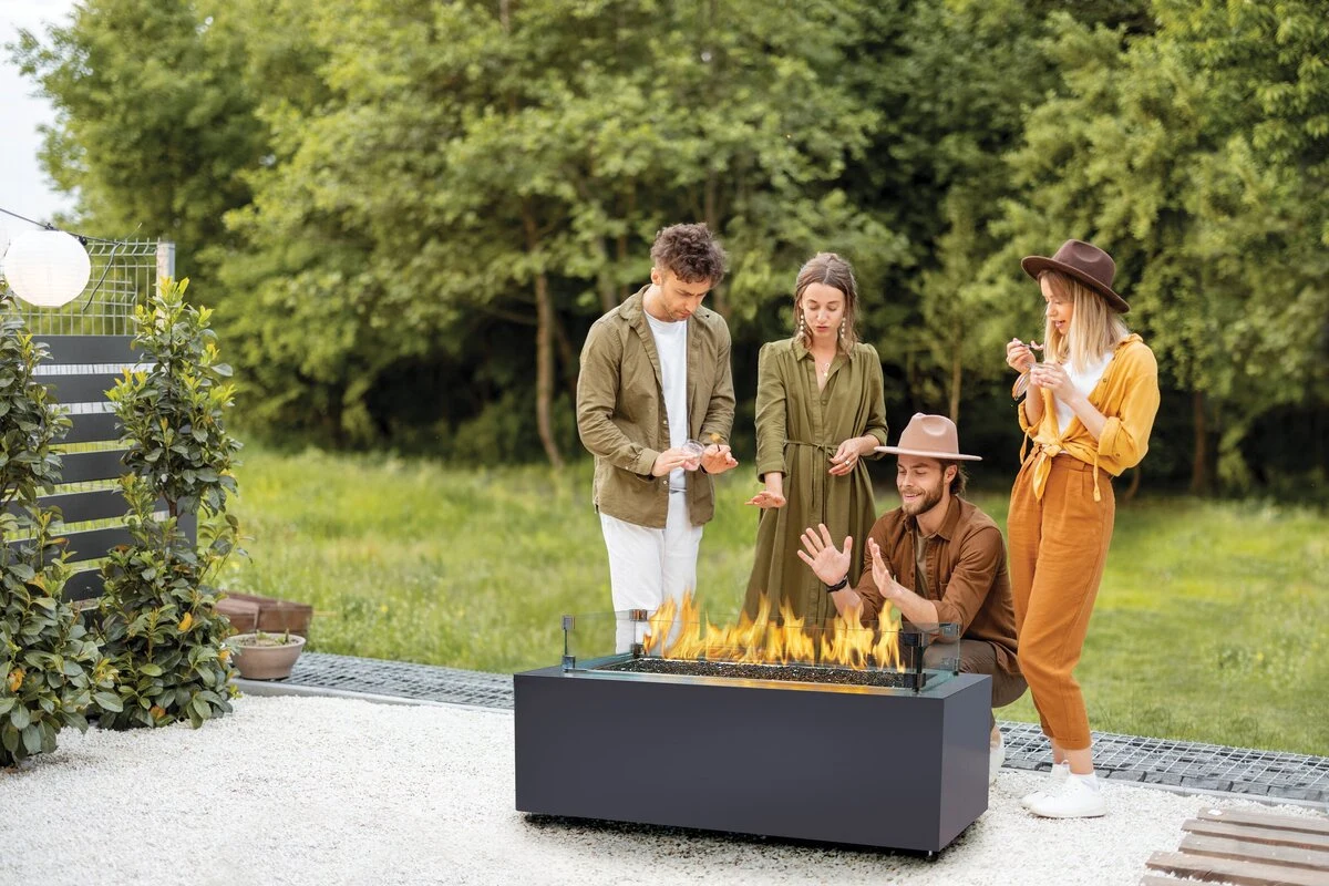 A family gathered around a rectangular concrete fire table with an active flame on a grassy lawn.
