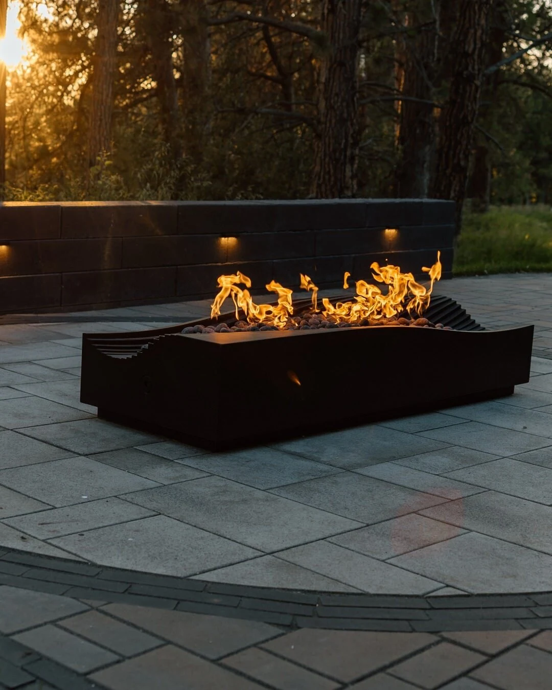 Modern rectangular gas fire table with flames burning on a paved patio at dusk.