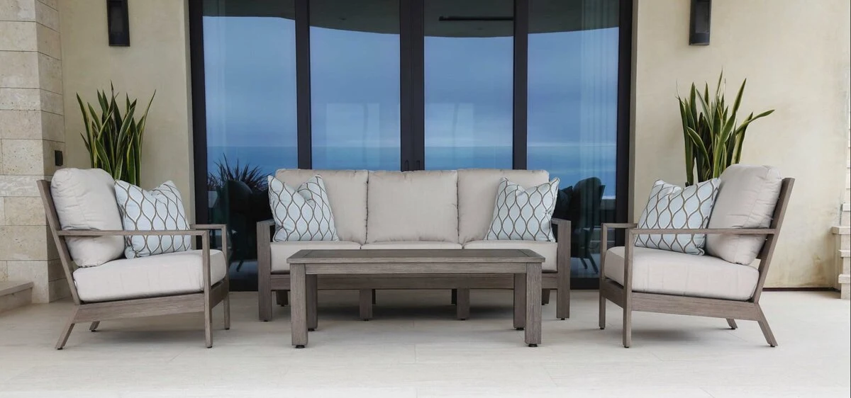 A black aluminum outdoor sofa and lounge chair with white cushions on a covered oceanfront patio with a wood pergola.