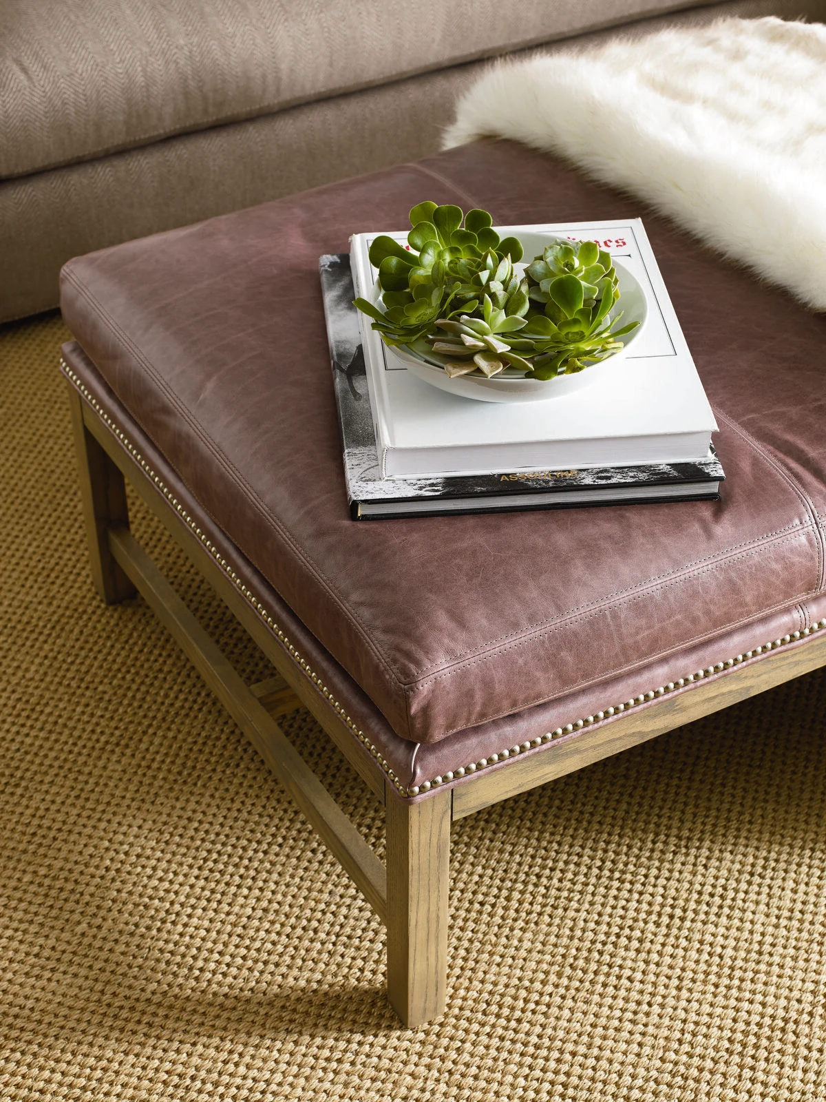 A close-up of a mauve leather nailhead ottoman with a stack of coffee table books and a succulent arrangement on top, set on a woven area rug.
