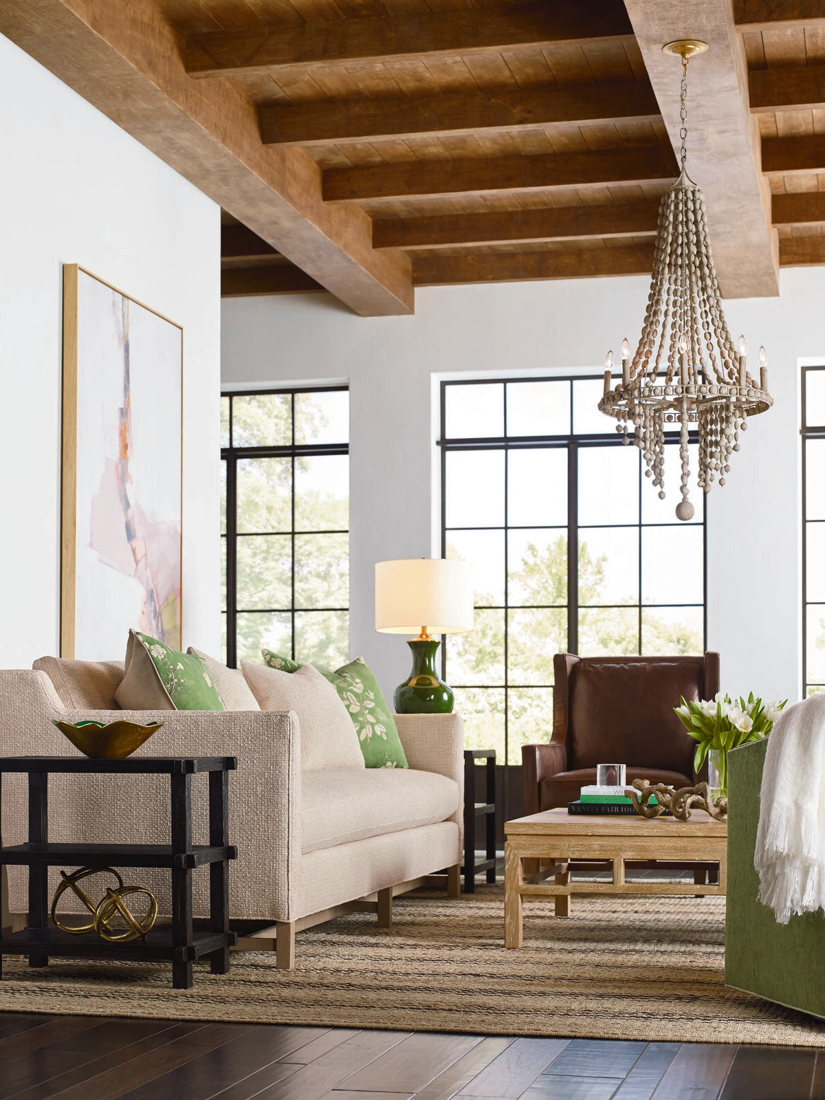 A bright open living room with white upholstered sofas, a dark wood coffee table, a beaded chandelier, exposed wood beam ceiling, black steel windows, and green accent pillows and plants.