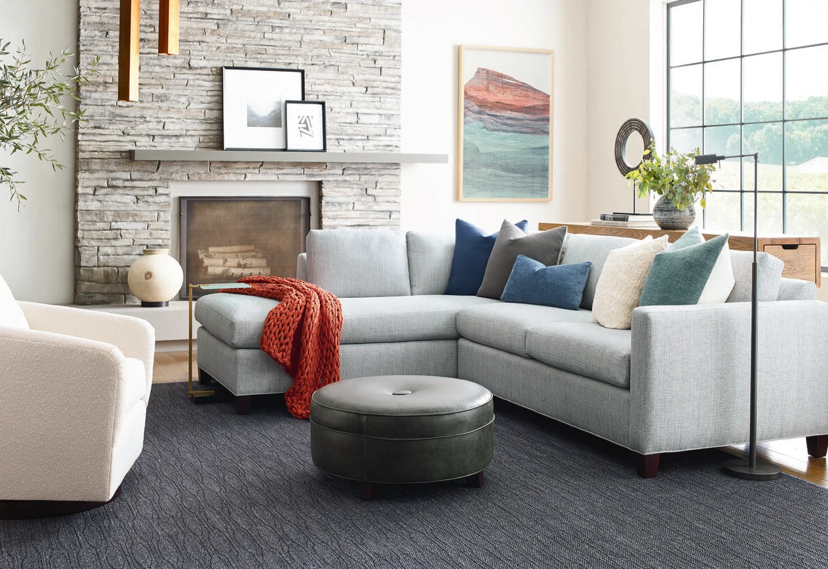 A light blue L-shaped sectional sofa with navy and teal throw pillows and a red throw blanket, paired with a dark green tufted round ottoman, in front of a stacked stone fireplace.