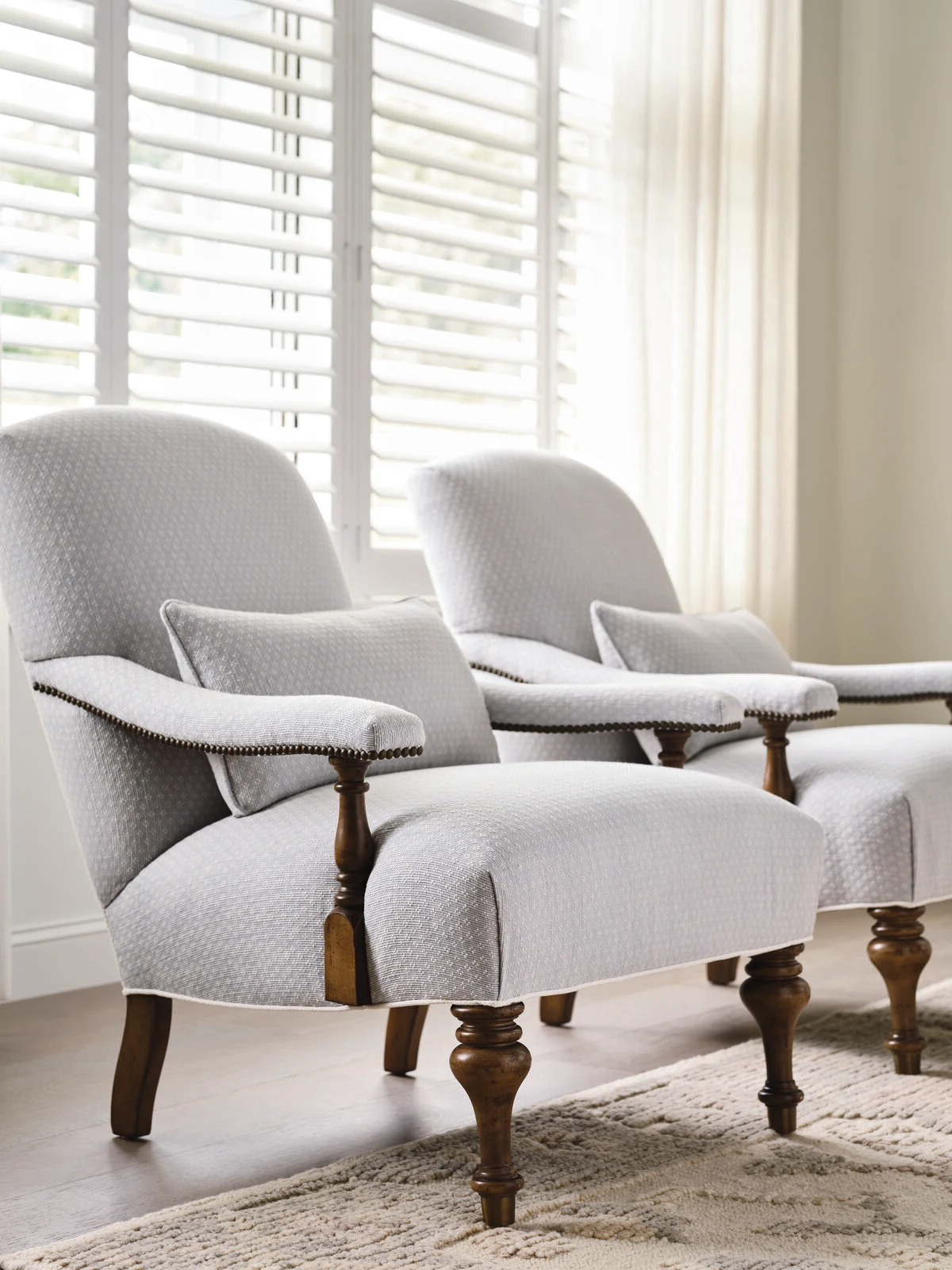 Two light blue upholstered accent chairs with dark wood carved legs on a neutral area rug in a bright room with plantation shutters.
