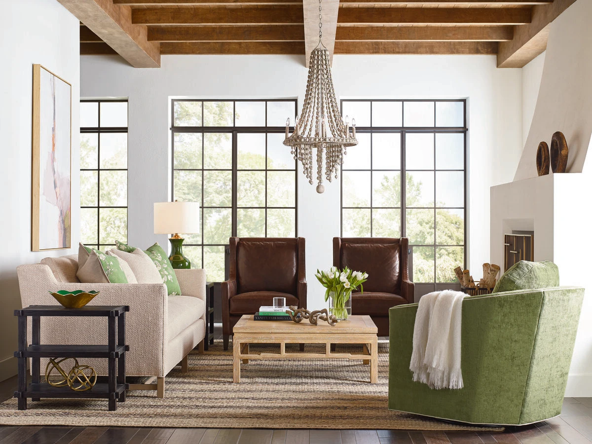 A living room with exposed wood beam ceilings, black steel windows, a beaded chandelier, a white upholstered sofa, dark brown leather chairs, green accent chairs, and green throw pillows on a neutral area rug.