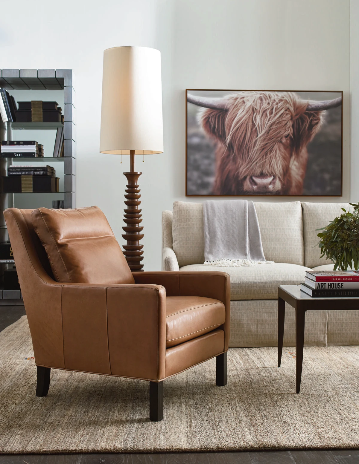 A living room with a caramel leather accent chair, a dark wood spindle floor lamp, a light gray upholstered sofa, and a large Highland cow canvas print above a side table with books.