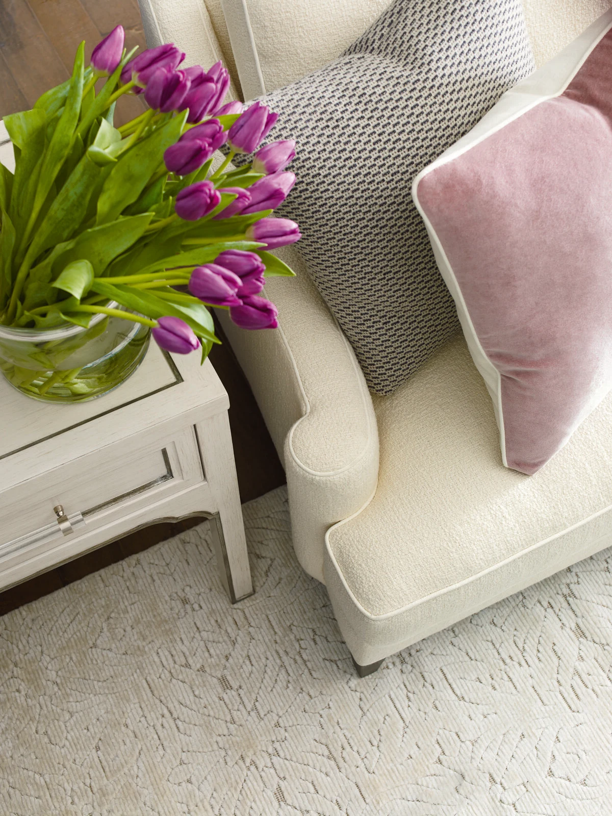 A close-up of a cream upholstered accent chair with a pink velvet throw pillow, next to a white side table with a vase of purple tulips on a light patterned area rug.