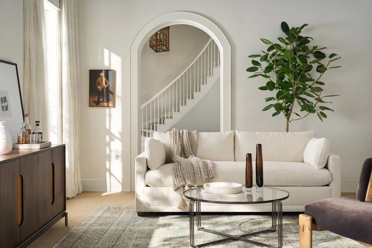 A bright minimalist living room with a cream upholstered sofa, round glass and metal coffee table, dark wood vases, a large fiddle-leaf fig tree, and a white arched entryway with a staircase beyond.