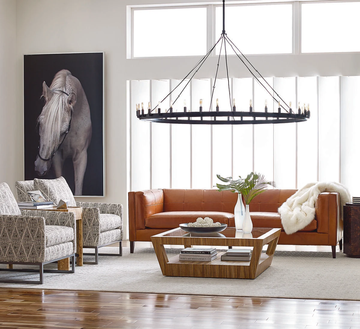 A contemporary living room with a rust orange leather sofa, patterned accent chairs, a wood and glass coffee table, a large dark horse portrait, and a black iron ring chandelier above large windows.