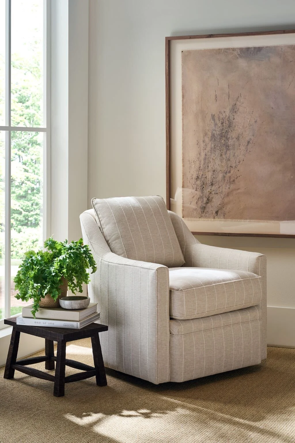 A bright corner vignette with a cream upholstered swivel accent chair, a dark wood stacked side table with a potted plant, and a soft abstract artwork on a warm plaster wall.