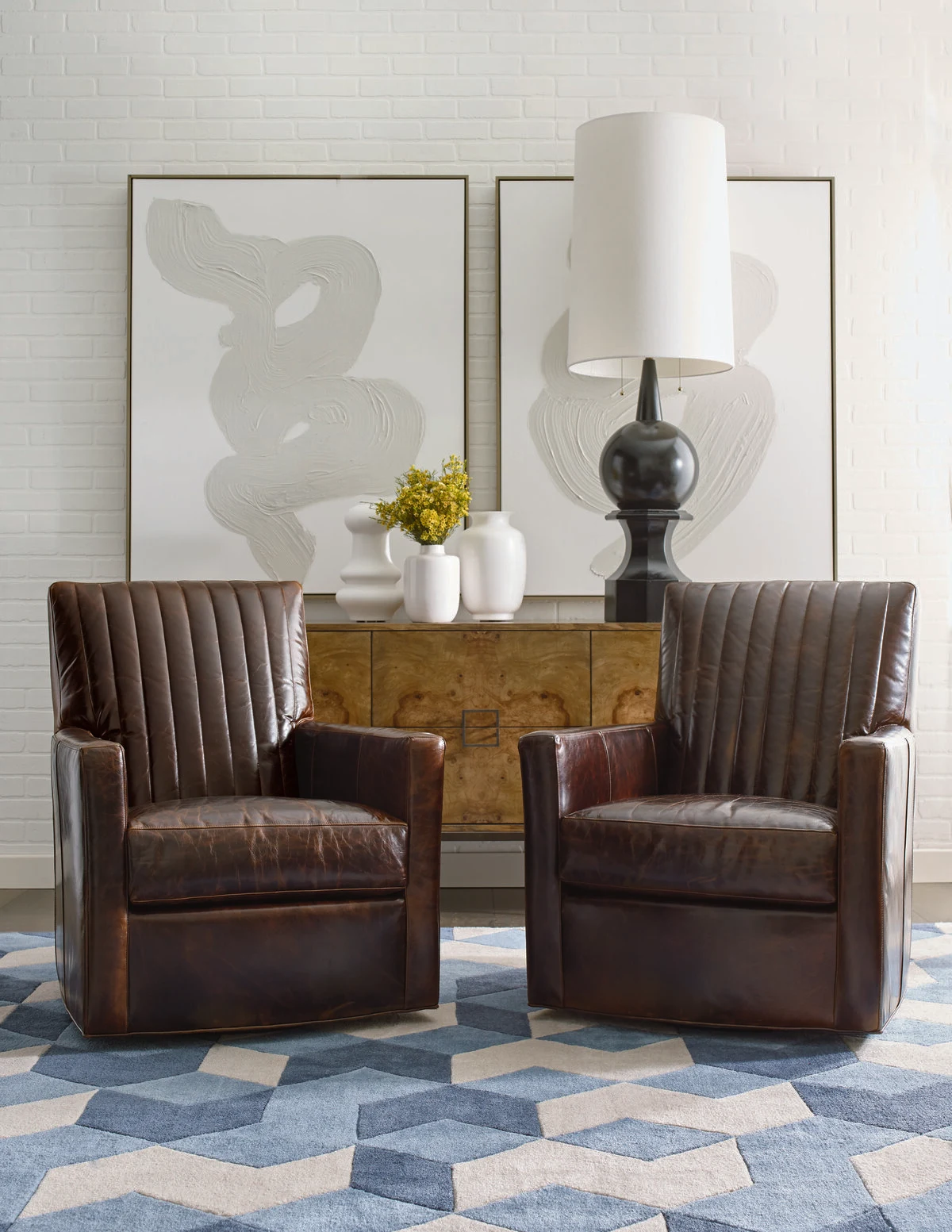 Two dark brown leather channel-back swivel accent chairs on a geometric blue area rug, with a gold burl wood credenza, abstract wall art, and dark ceramic vases behind them.