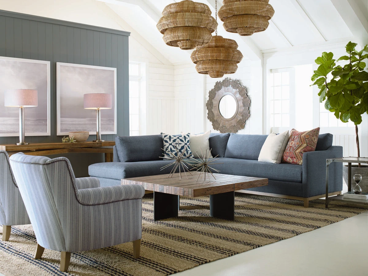A teal and blue living room with a blue upholstered sofa and striped accent chair, a wood and glass coffee table, a striped area rug, layered woven pendant lights, and a sunburst mirror on a teal accent wall.