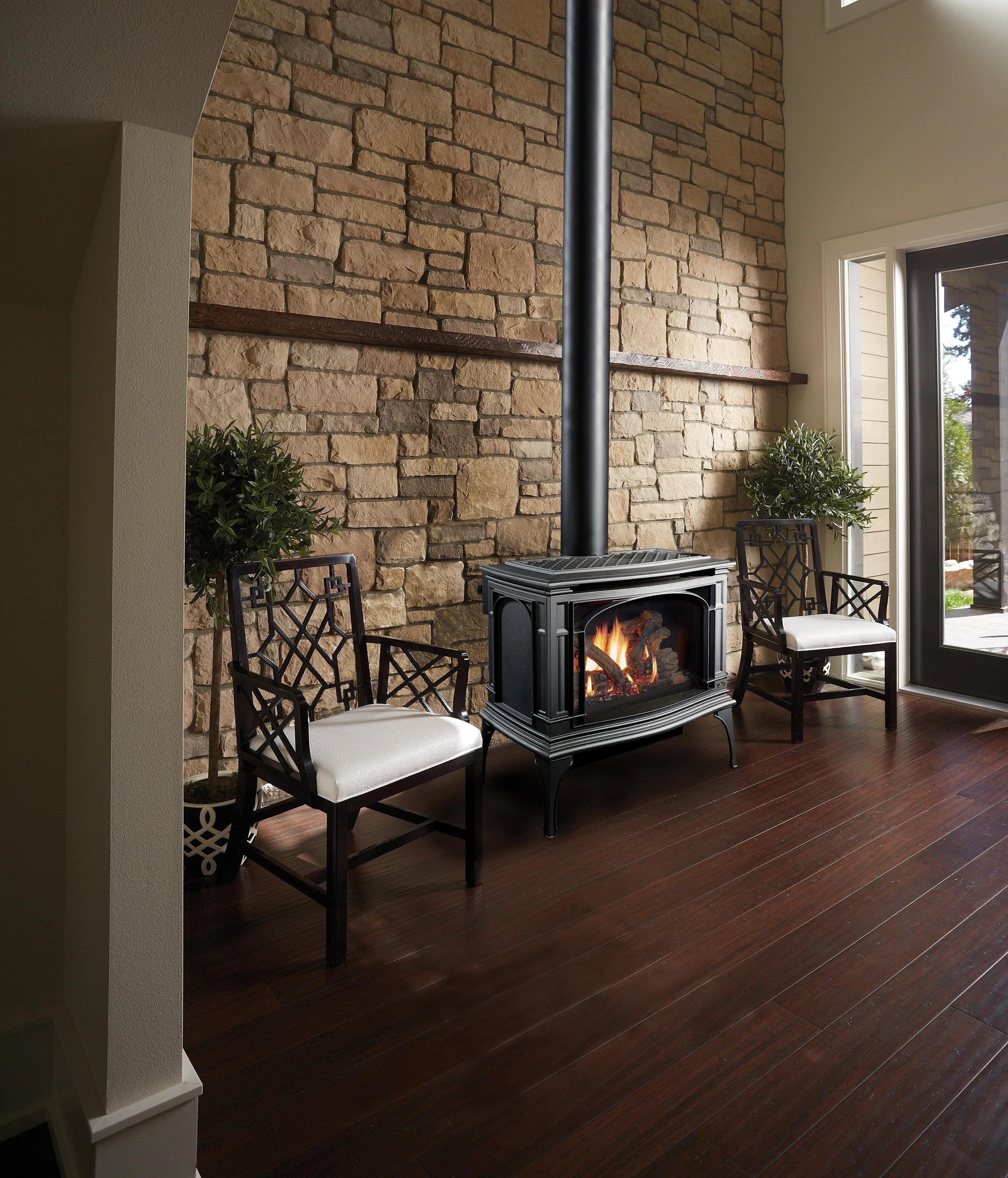 Lopi Greenfield Millivolt Gas Stove against a floor to ceiling stone accent wall with dark hardwood floors and white accent chairs nearby.