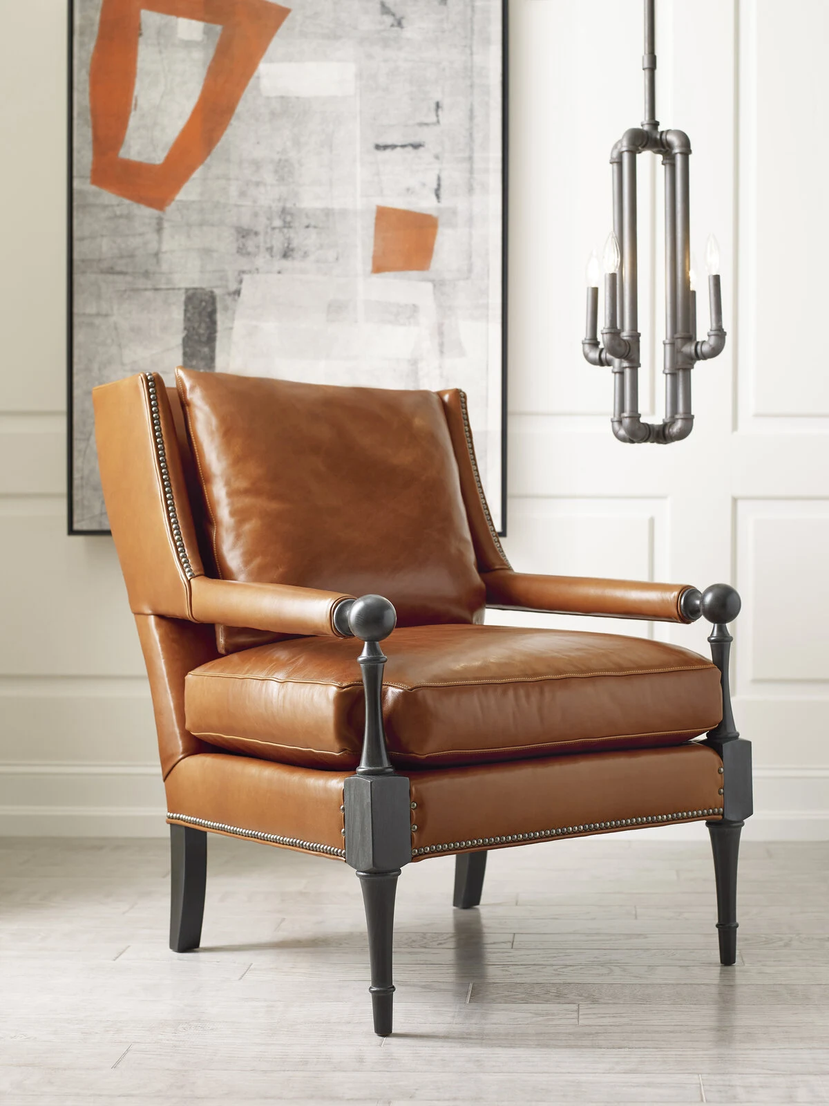 A cognac leather accent chair with nailhead trim and dark wood ball-and-post arms and legs, set against a white paneled wall with an abstract artwork and an iron candelabra sconce.
