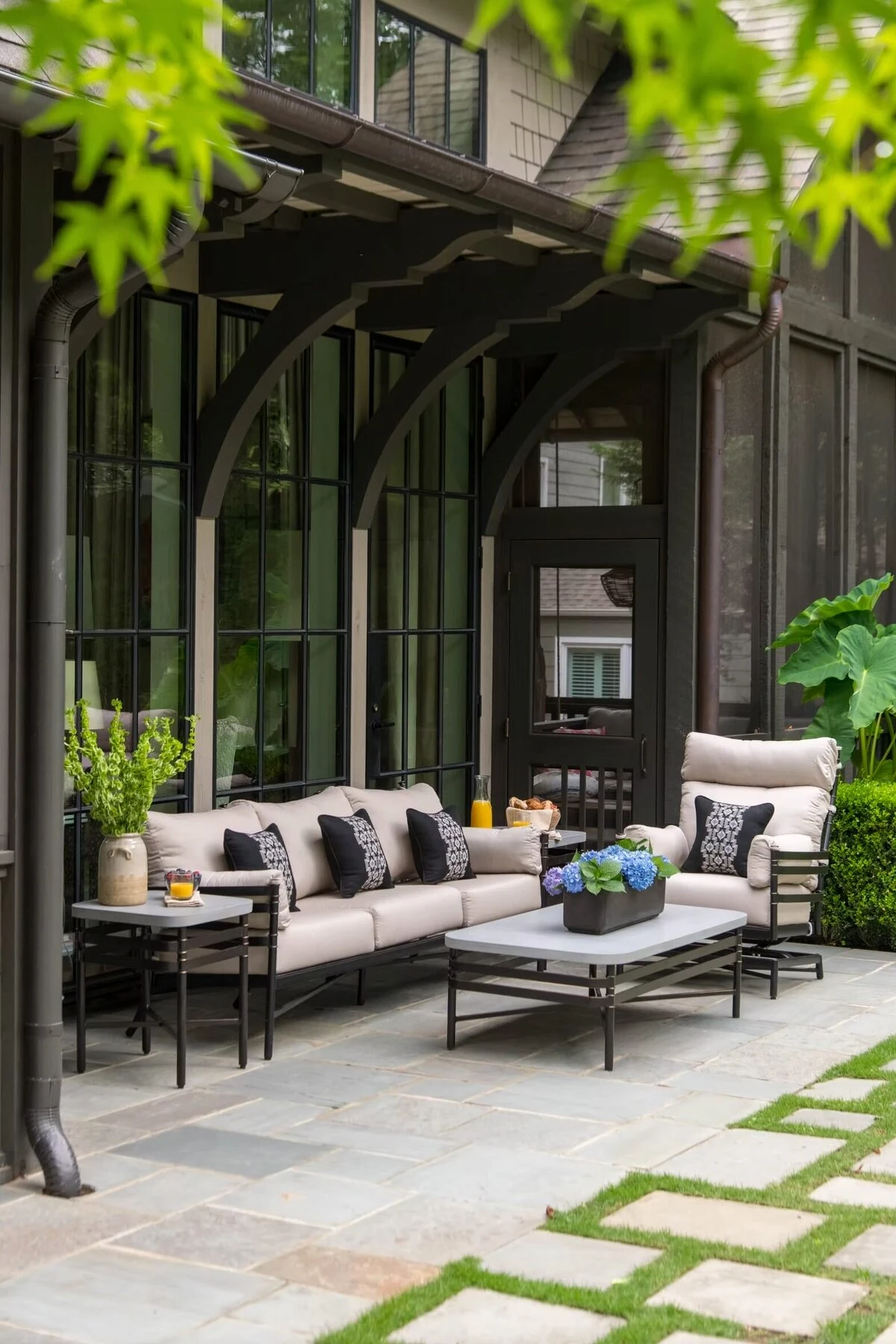 A dark aluminum outdoor sectional sofa and lounge chairs with neutral cushions and decorative pillows on a stone patio with large arched black steel windows and lush landscaping.