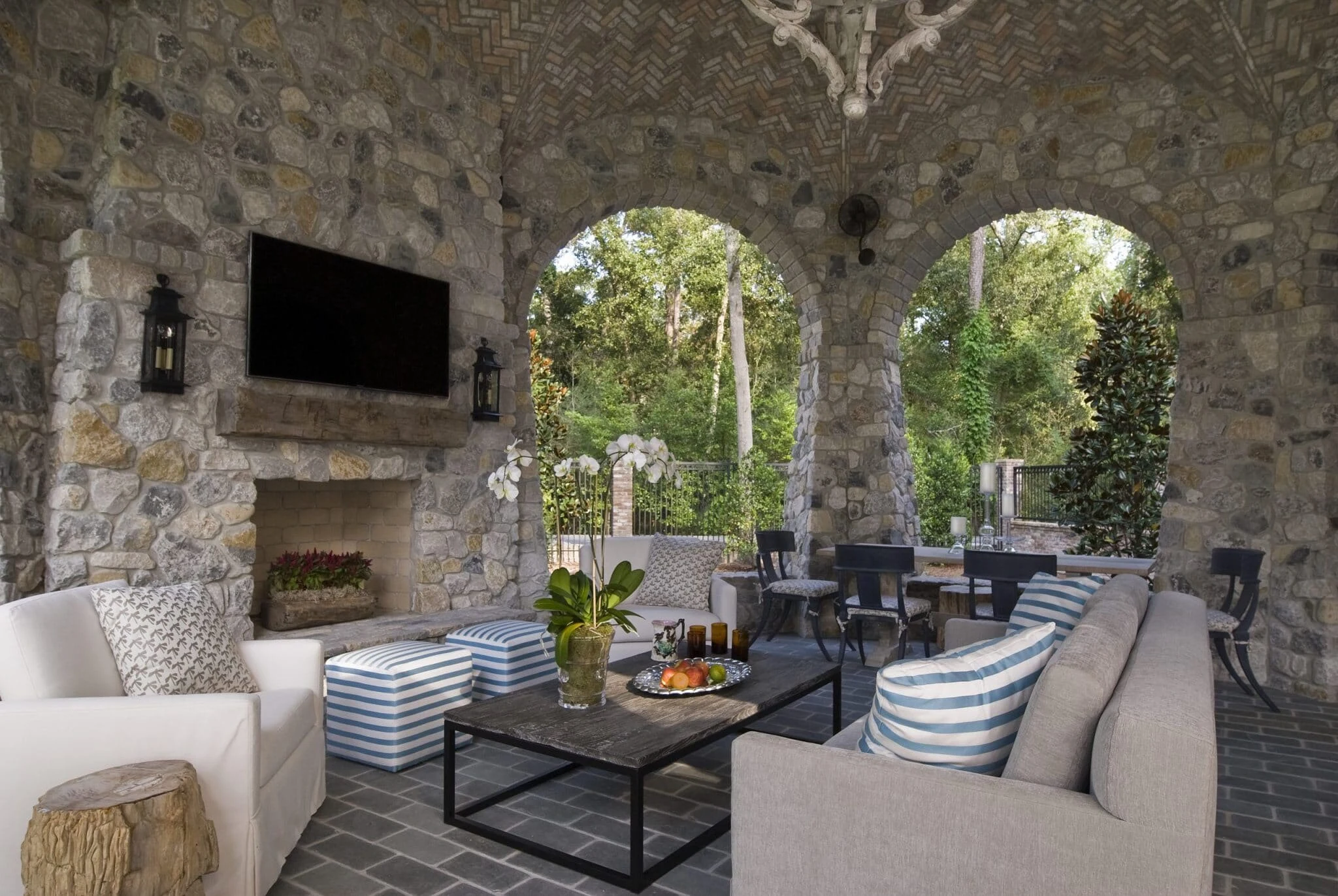 A masonry fireplace set into a stone exterior wall of a covered outdoor living space surrounded by mature trees and modern furniture.