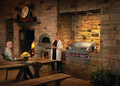 Napoleon Built-In 700 Series 38 grill installed in a stone outdoor kitchen beside a wood-fired pizza oven.