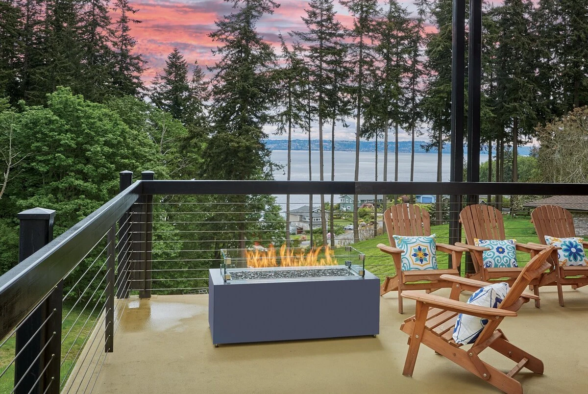 A rectangular linear fire table with an active flame on a deck overlooking a lake and forested landscape.