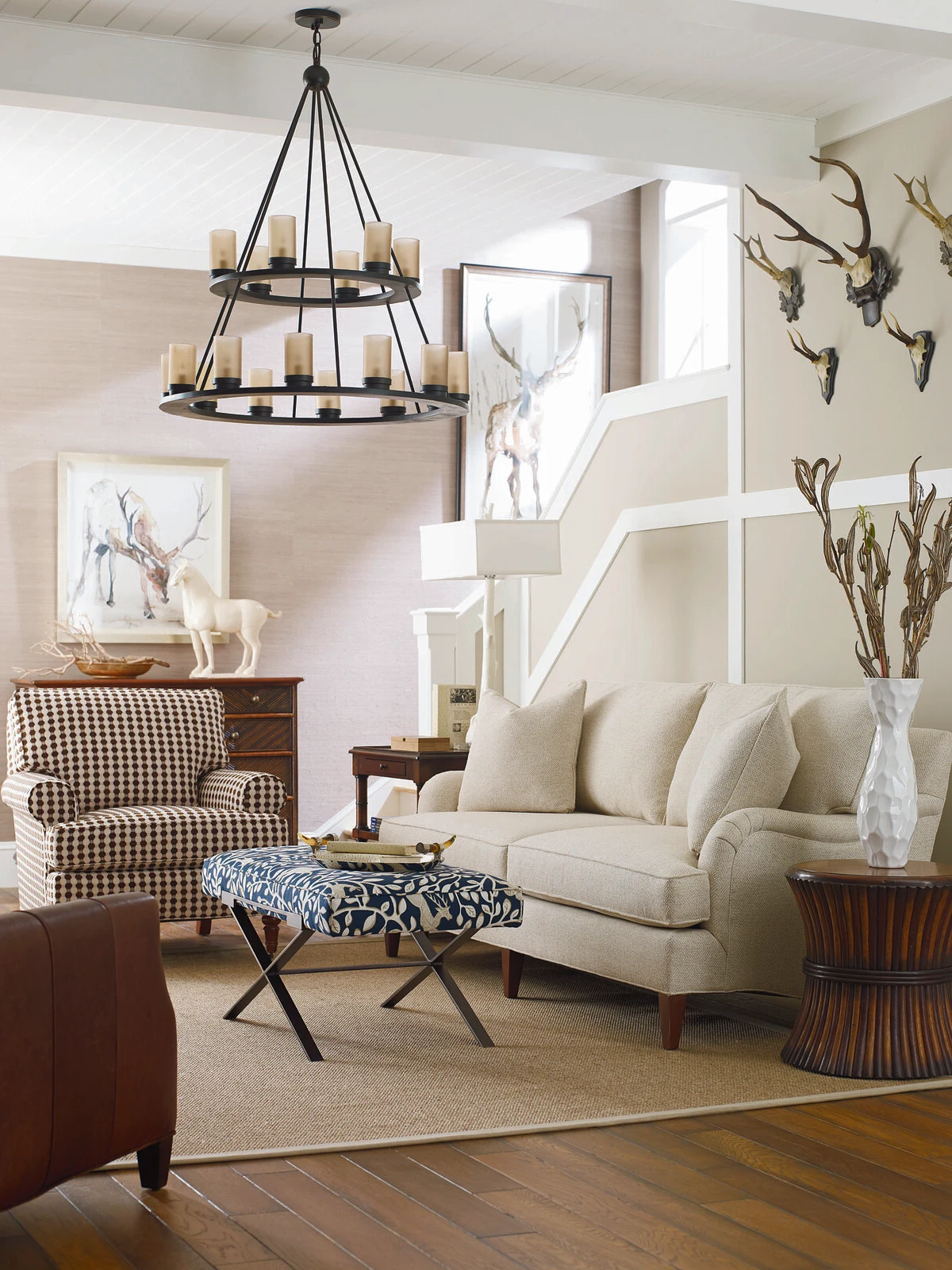 A transitional living room with a cream upholstered sofa, a houndstooth accent chair, a blue patterned bench, dark wood side tables, an iron candle chandelier, and antler wall decor on a sage green accent wall.