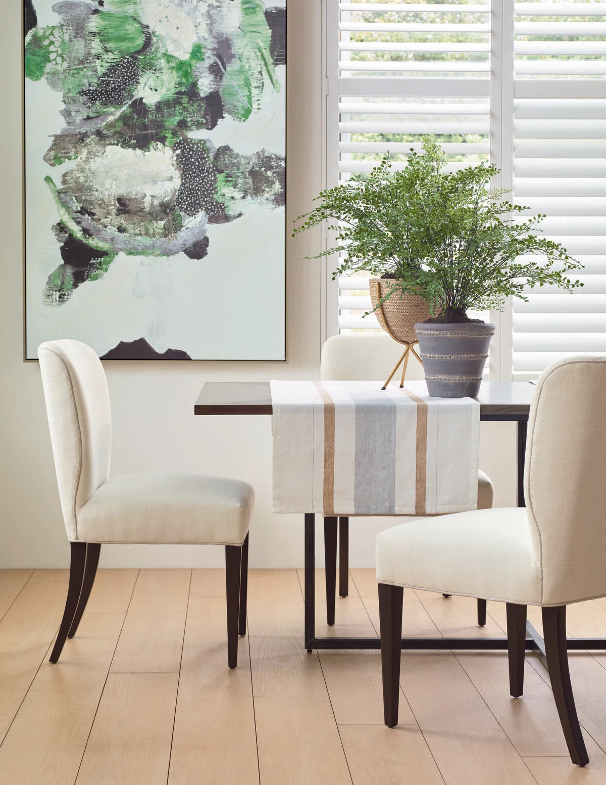 A bright dining area with cream upholstered dining chairs and a white and dark wood dining table, a large green abstract artwork, a potted fern, and plantation shutters.