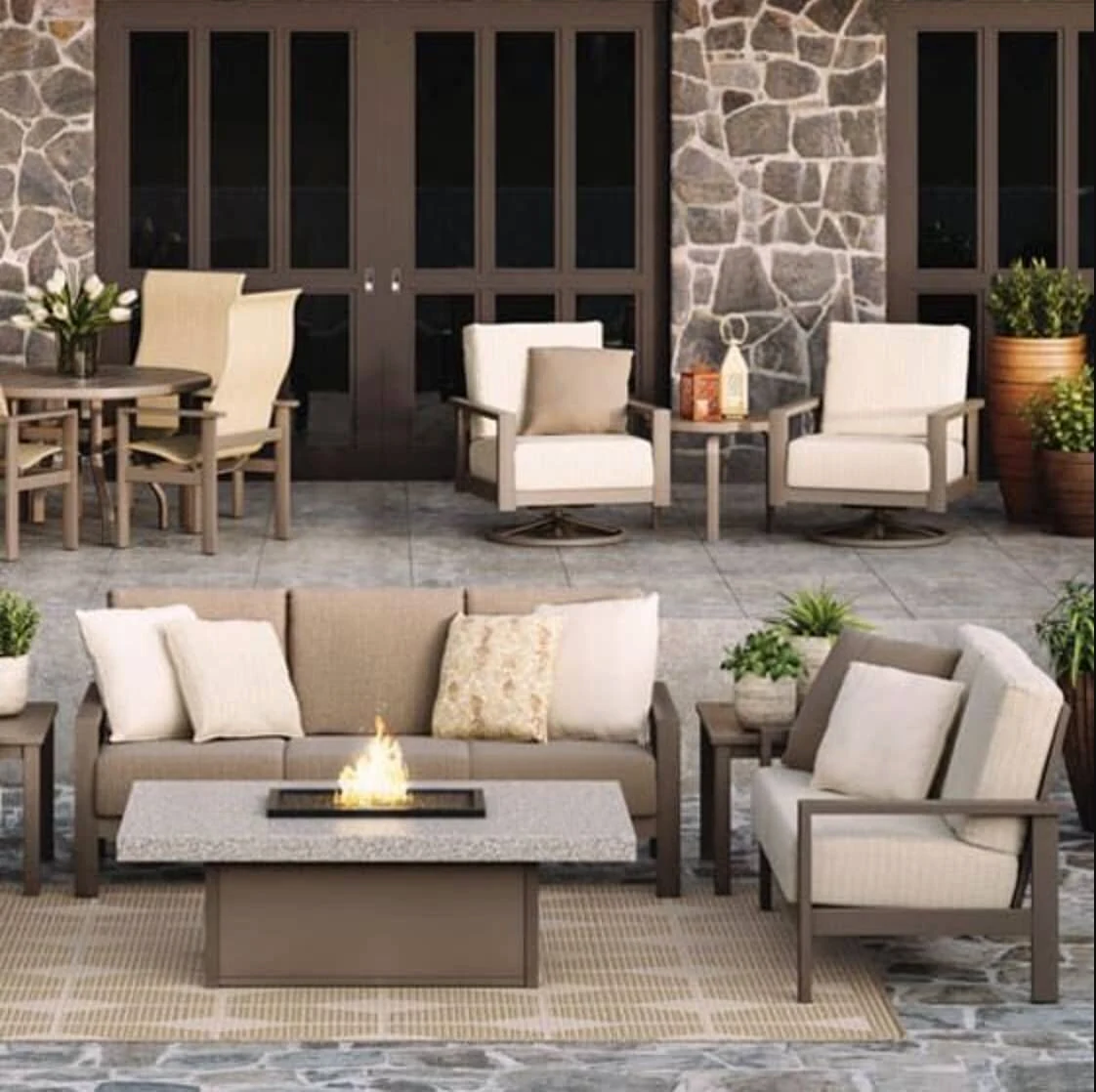 A gray aluminum outdoor sofa and swivel lounge chairs with neutral cushions arranged around a rectangular fire table on a stone patio with a stacked stone wall backdrop.