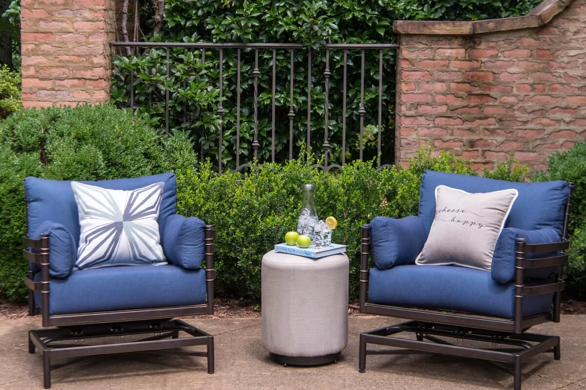 A dark aluminum outdoor sofa and lounge chairs with blue cushions and decorative pillows arranged around a round upholstered ottoman on a brick patio with iron fencing and landscaping.