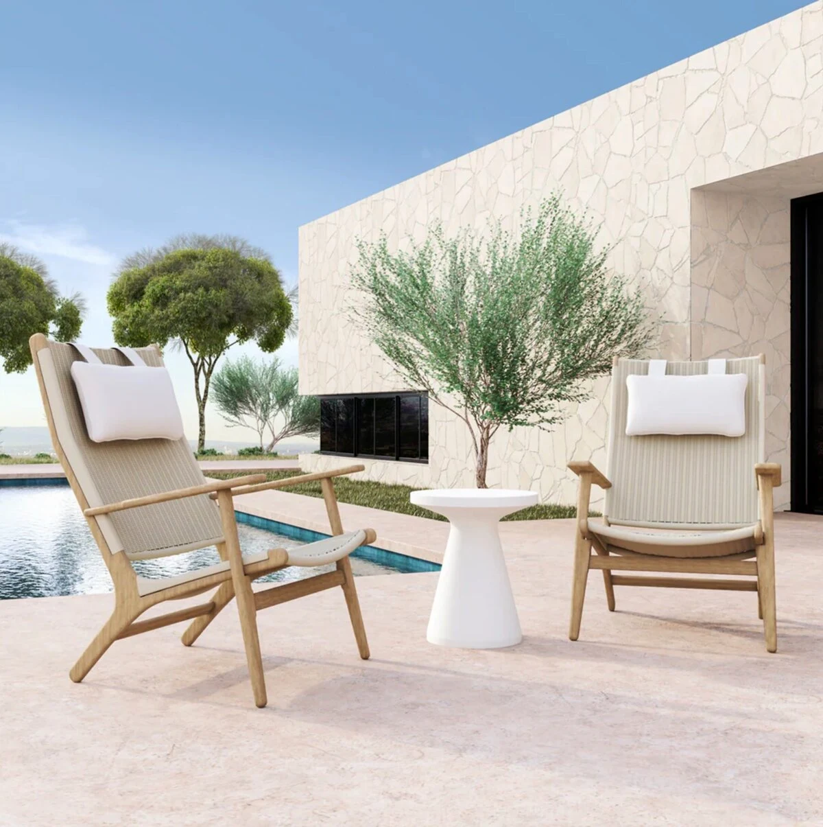 Two teak high-back lounge chairs with neutral cushions and a round white side table on a light stone patio beside a pool, with an olive tree and modern white stucco exterior in the background.