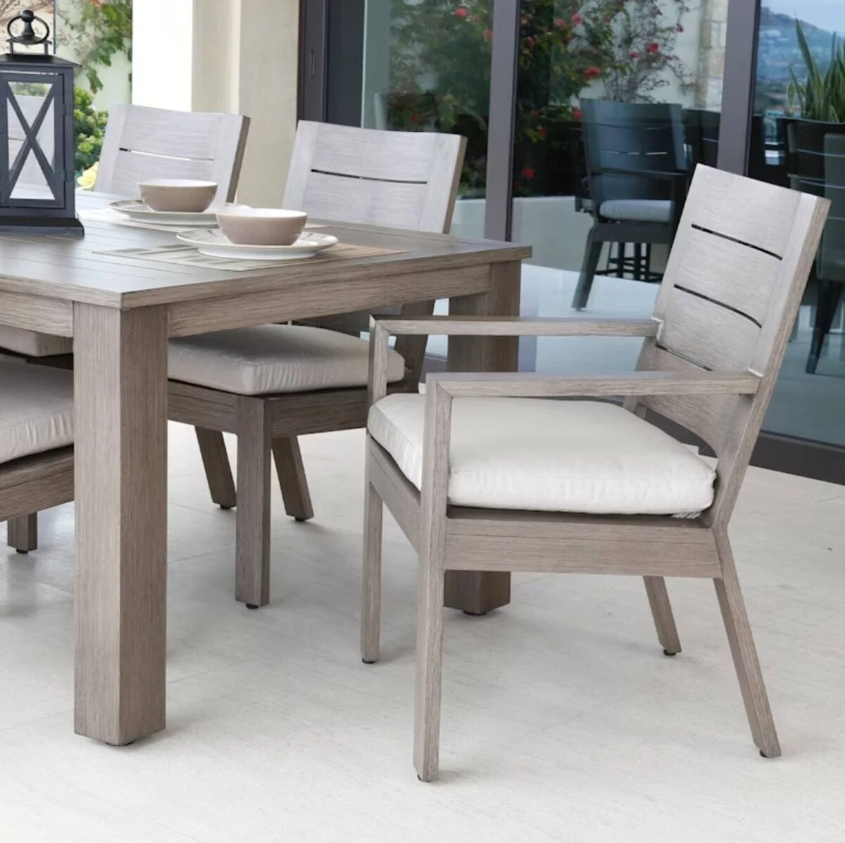 A weathered teak outdoor dining table and cushioned dining chairs with neutral cushions on a patio with large windows and a lantern accent.