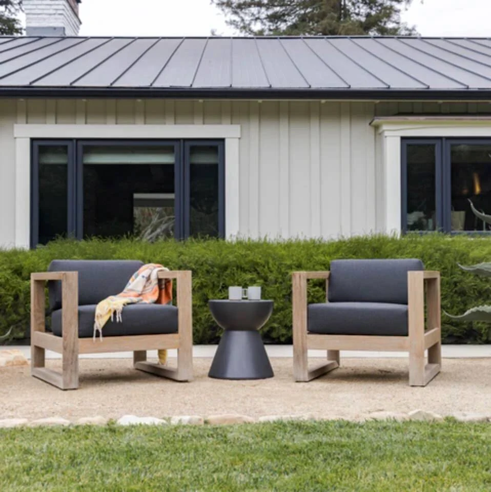 Two teak outdoor lounge chairs with dark gray cushions and a round dark metal side table on a lawn in front of a modern white and gray home exterior.