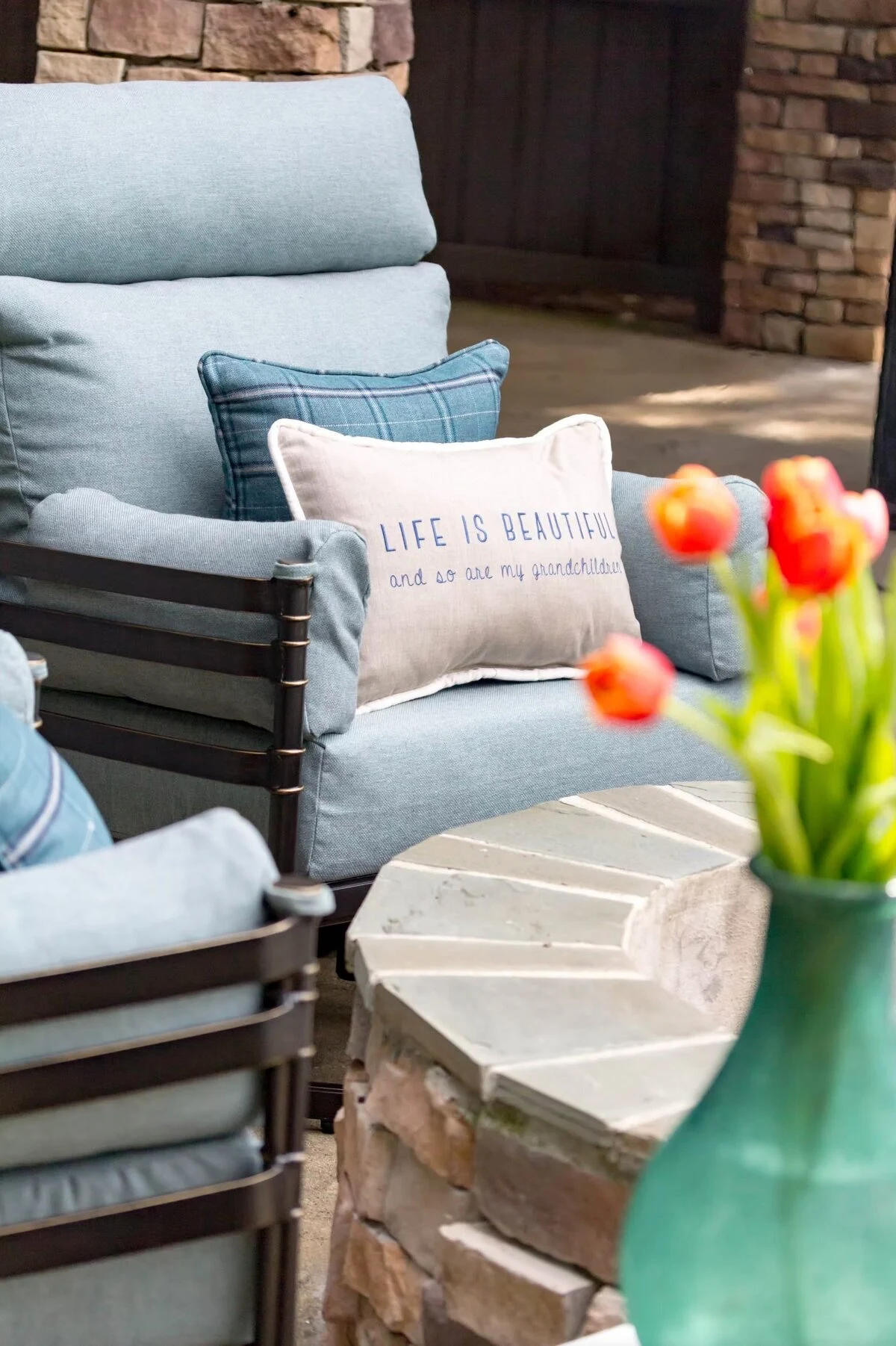 A close-up of a dark aluminum outdoor lounge chair with blue cushions and decorative pillows, including a "Life is Beautiful" accent pillow, with a vase of red tulips nearby.