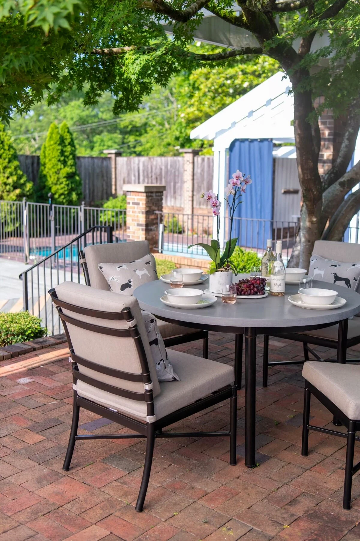 A dark aluminum outdoor round dining table set with dishes and a centerpiece, surrounded by cushioned dining chairs on a brick patio beside a pool with lush landscaping.