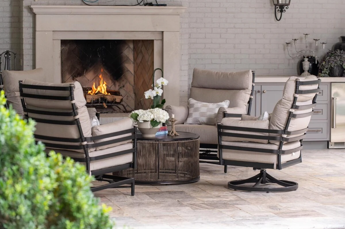 Two dark aluminum swivel lounge chairs with neutral cushions arranged around a round side table in front of a white brick outdoor fireplace with an active fire.