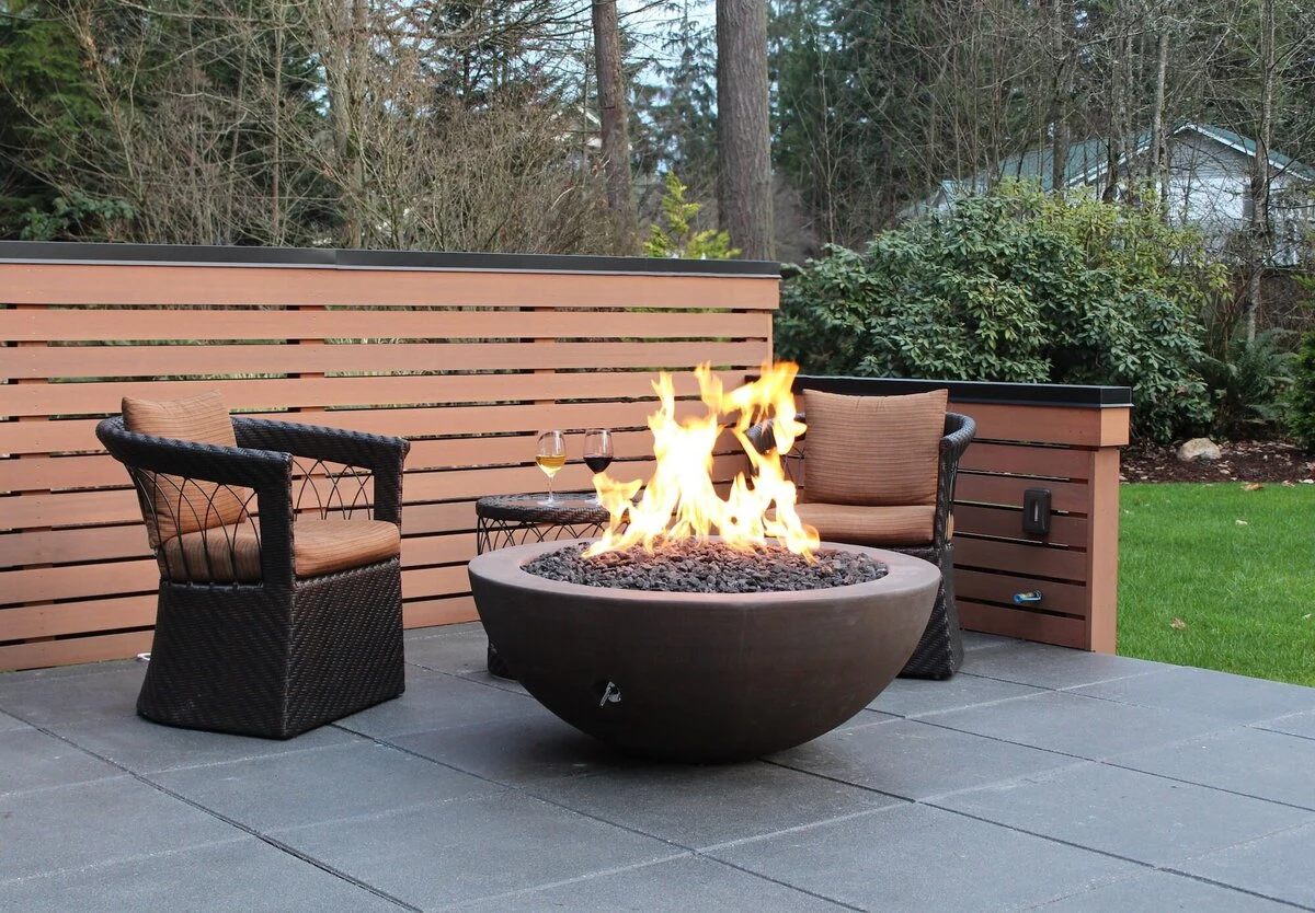 A round concrete fire bowl with an active flame on a wood deck surrounded by outdoor seating.