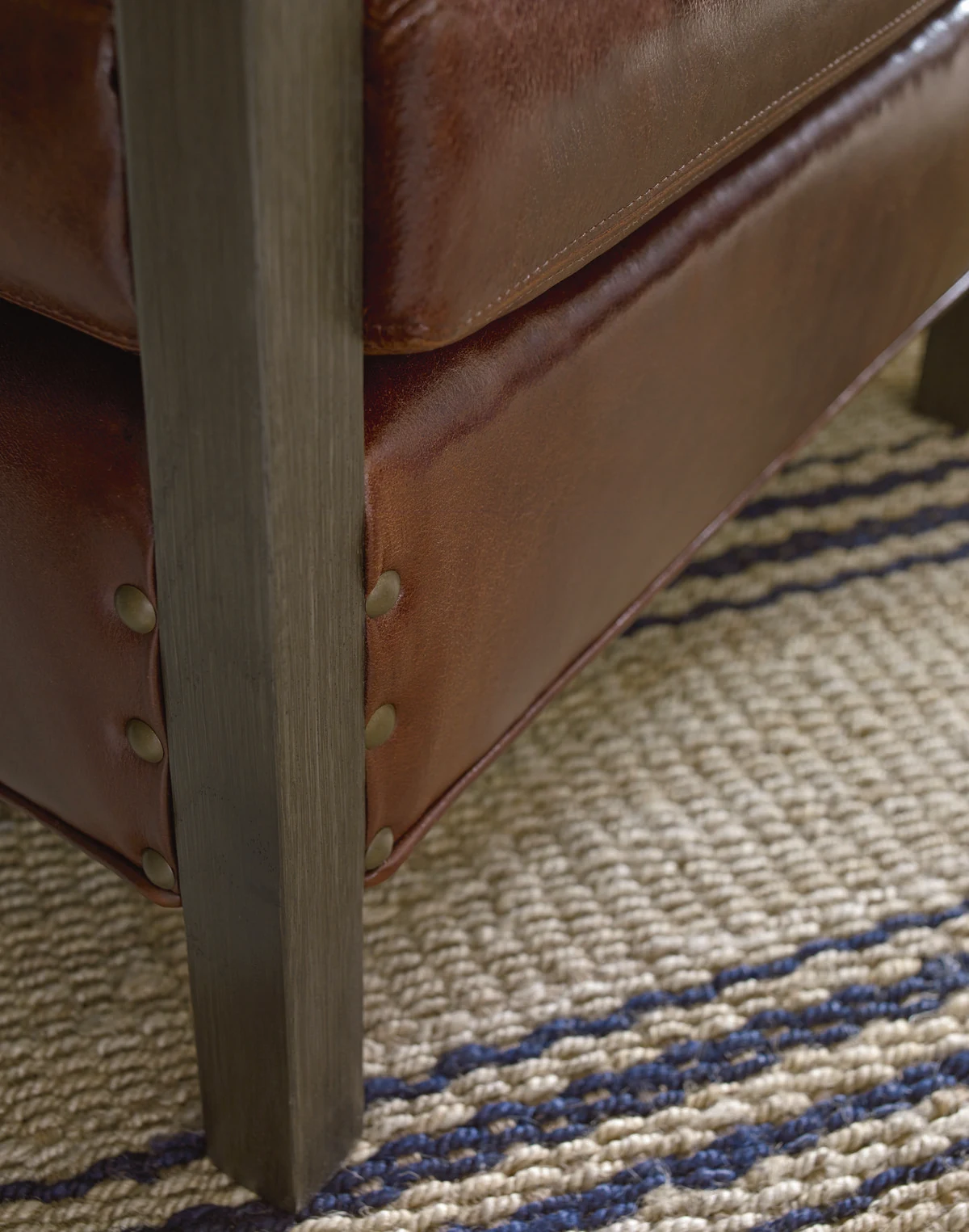A close-up of a dark wood furniture leg with nailhead trim detail on a cream upholstered piece, set on a blue and white woven area rug.